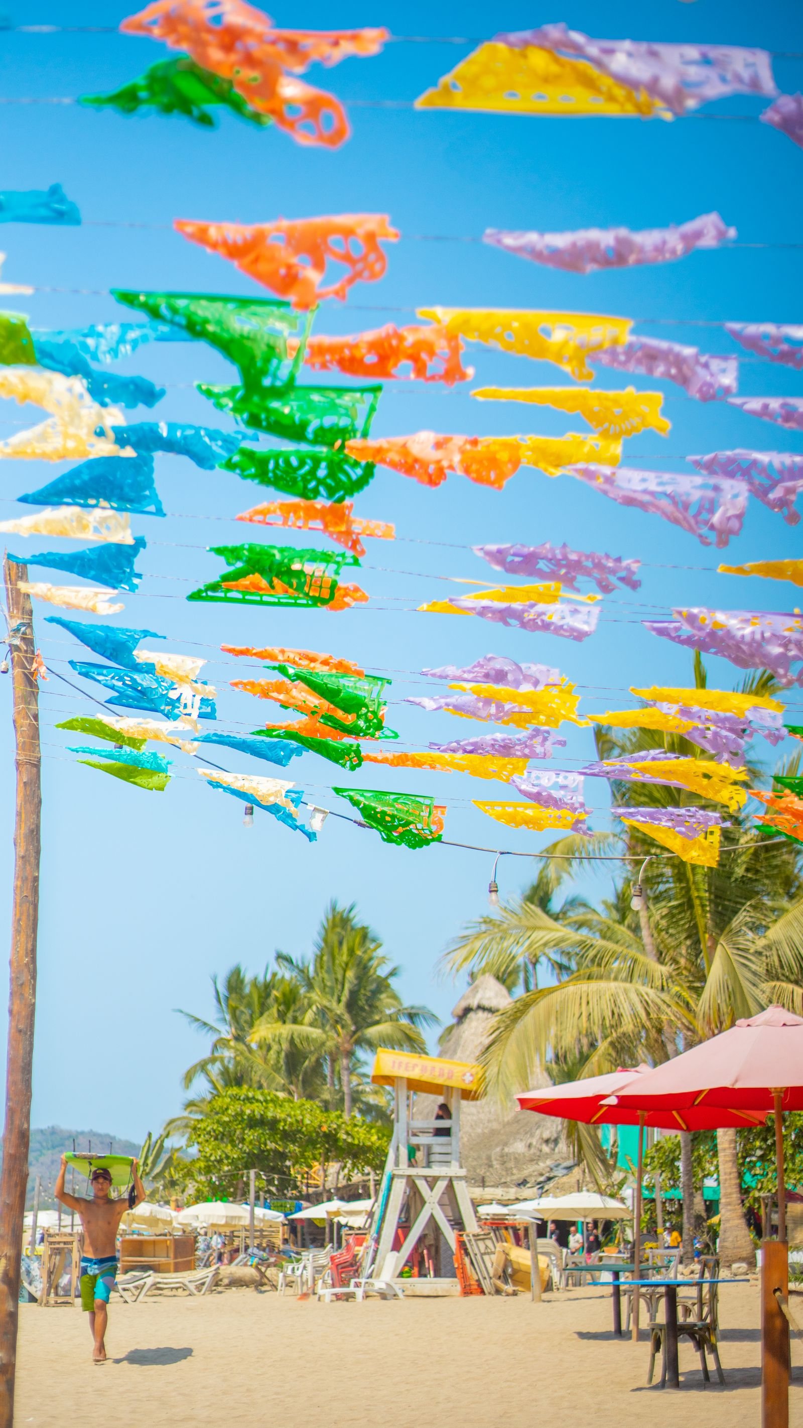 Colorful papel picado over Sayulita beach, with a lifeguard tower and a surfer in the background