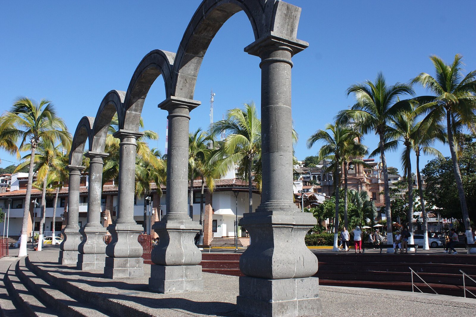 Four gray stone arches of the Los Arcos amphitheater on the Malecón, with palm trees and the church tower behind