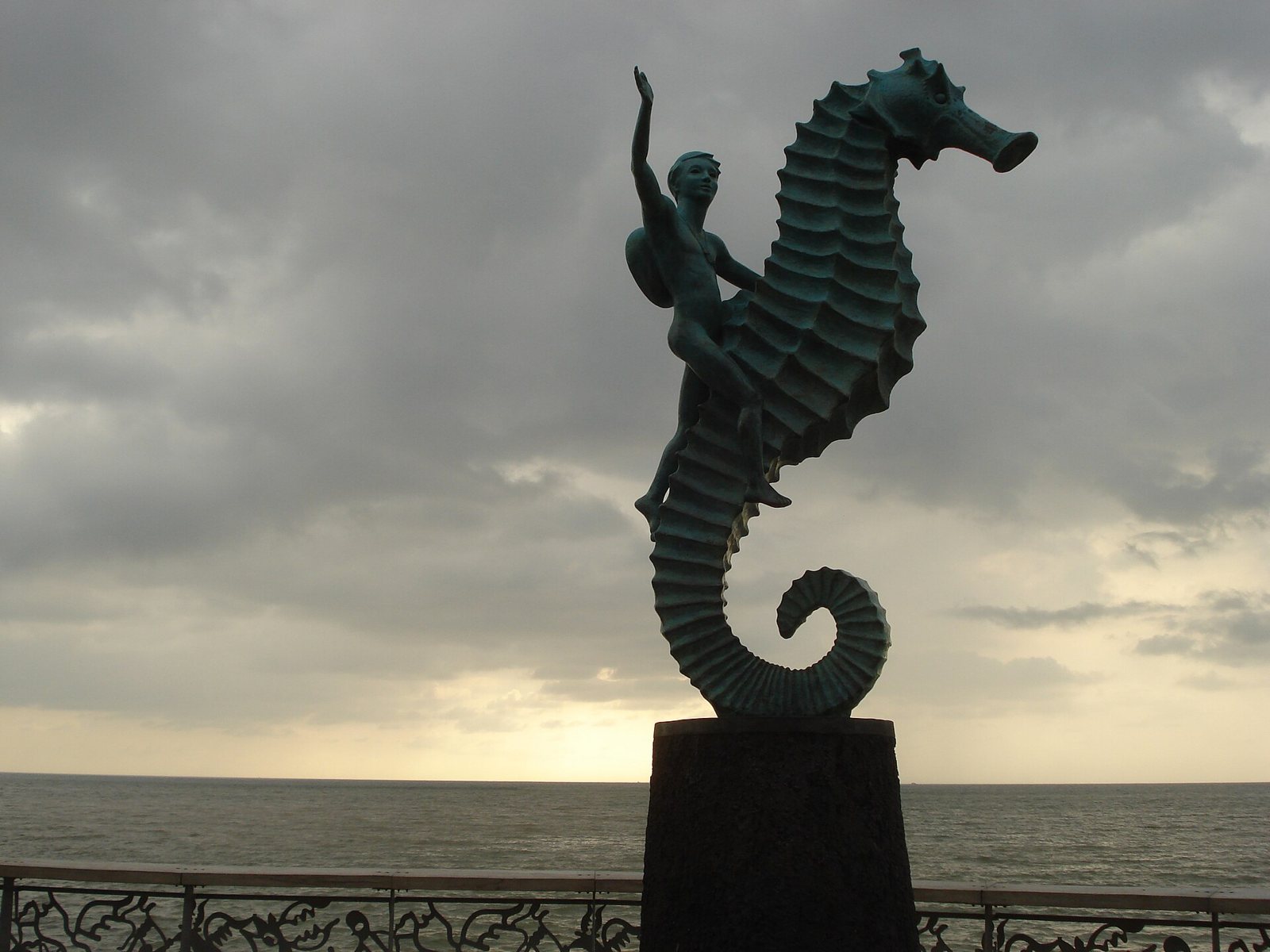 Bronze sculpture of a boy riding a seahorse on a pedestal, with the ocean and a cloudy sky behind