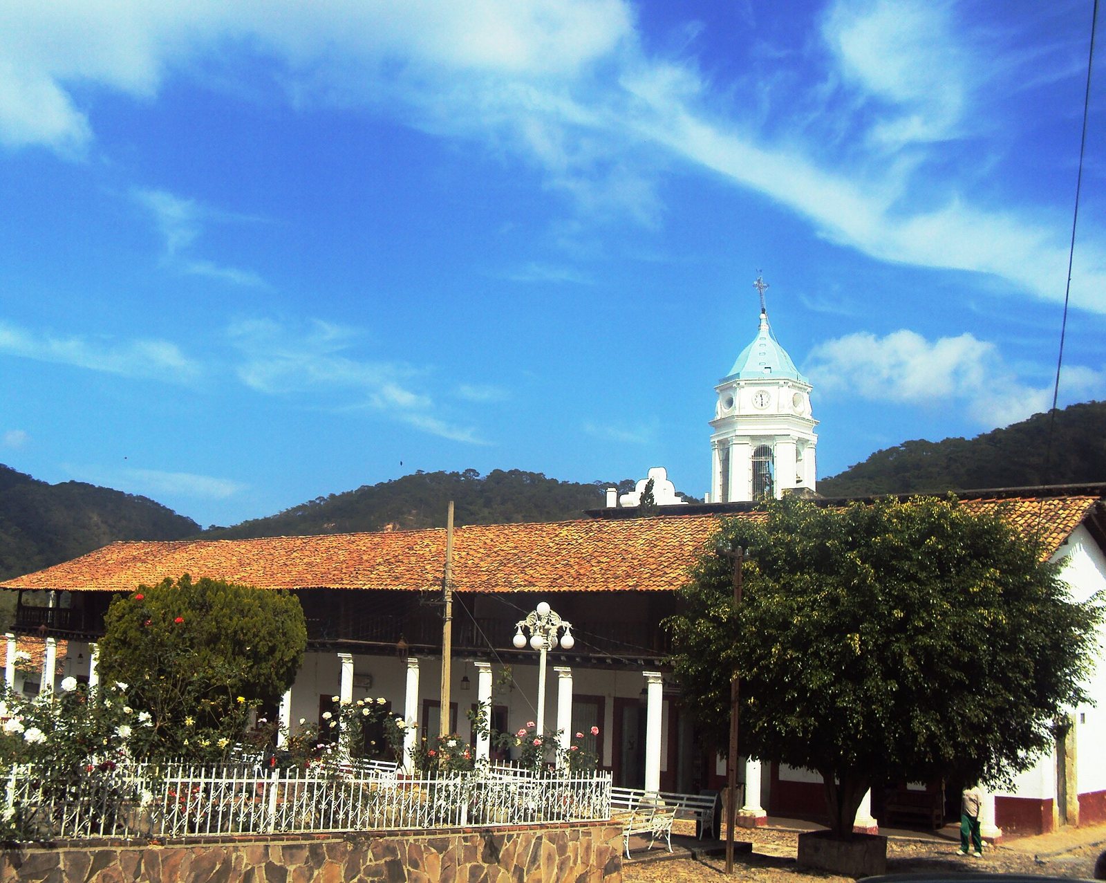 White bell tower of the San Sebastián church and tile-roofed arcades, with forested hills behind