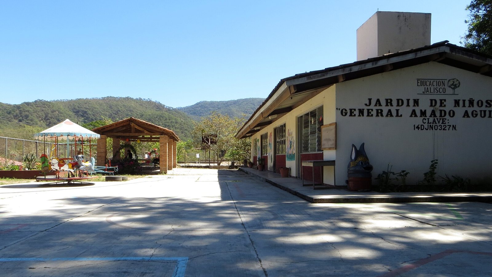 Kindergarten building with "General Amado Aguirre" painted on the facade, a schoolyard and forested mountains behind