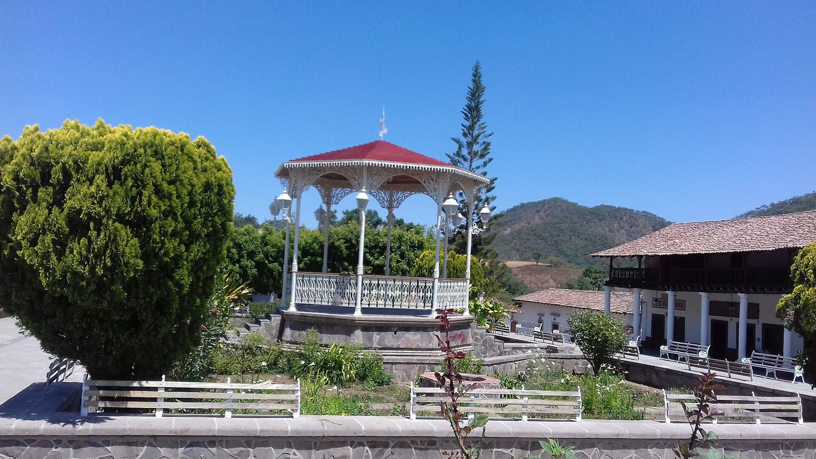 White bandstand with a red roof in the plaza of San Sebastián del Oeste, with gardens, benches and hills behind