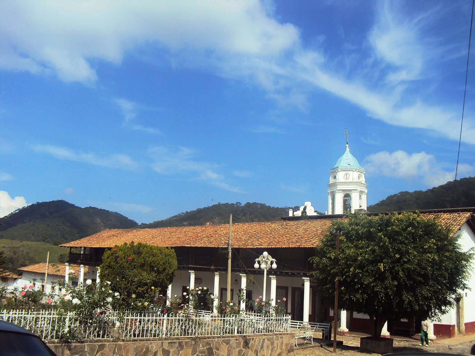 White bell tower of the San Sebastián church behind an arcaded building with a tile roof, a rose garden and forested hills behind