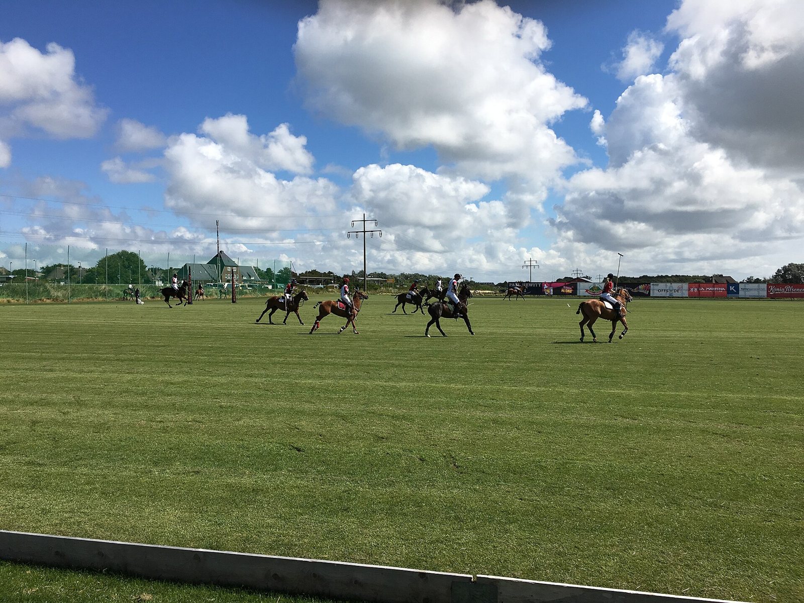Wide view of a polo field with several riders in play under a blue sky with big white clouds