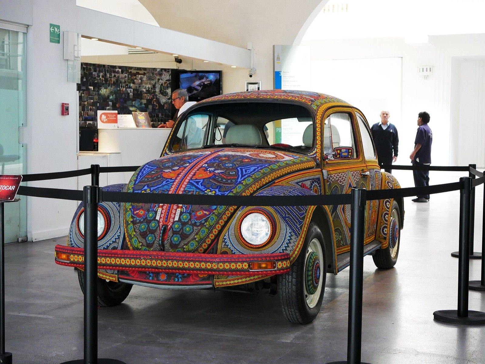 Volkswagen Beetle covered in beads with Huichol-style designs, displayed behind a rope barrier in a museum lobby