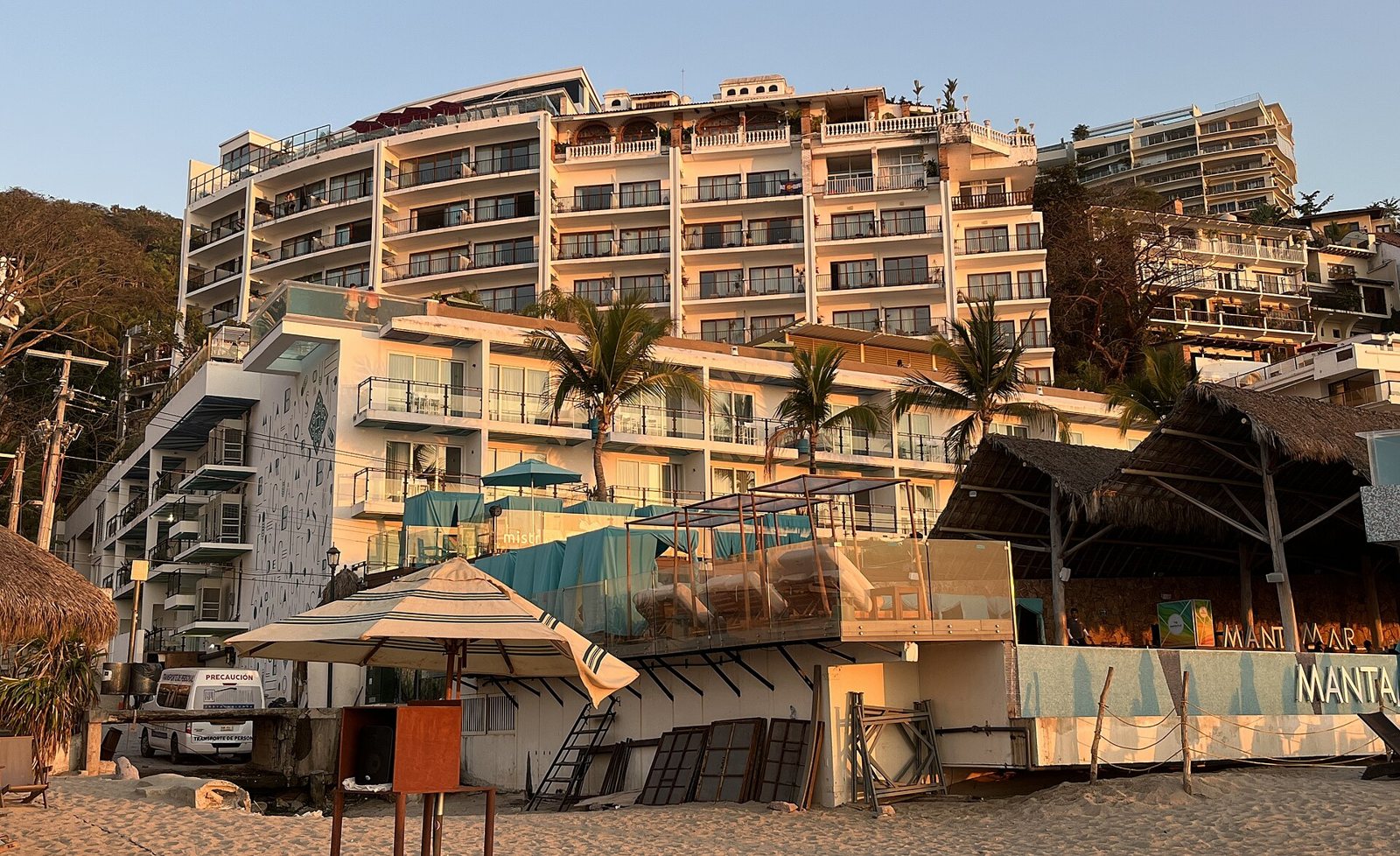 White towers of the Almar hotel with balconies above its beach club with turquoise cabanas on the sand of Los Muertos Beach, in golden evening light