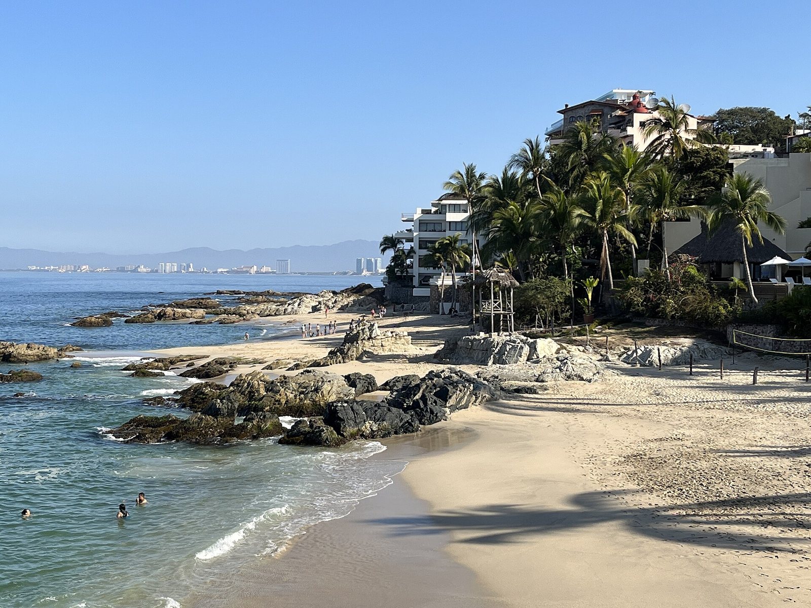 Light sand beach with dark rocks, palm trees and hillside buildings, with the Banderas Bay coastline in the distance