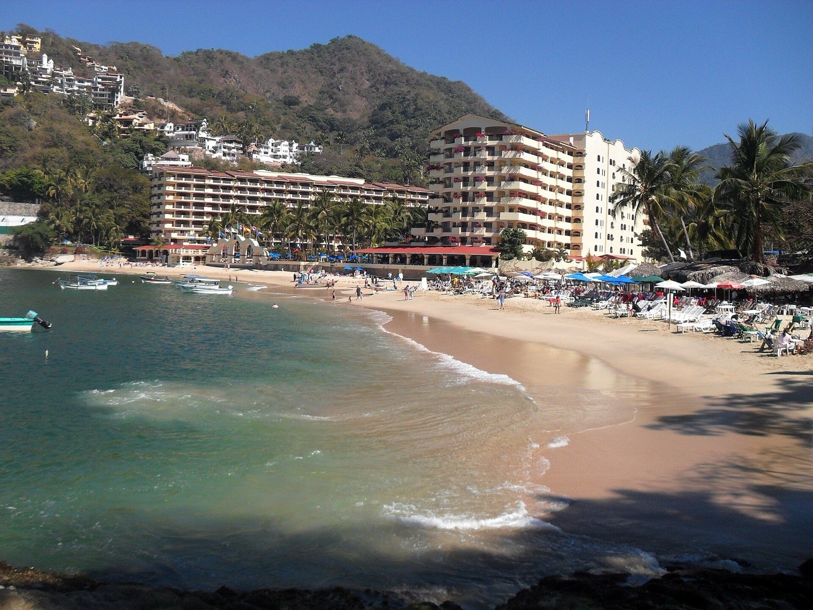 Crescent-shaped Mismaloya Beach with boats, palapas, beachfront hotel buildings and green hills behind