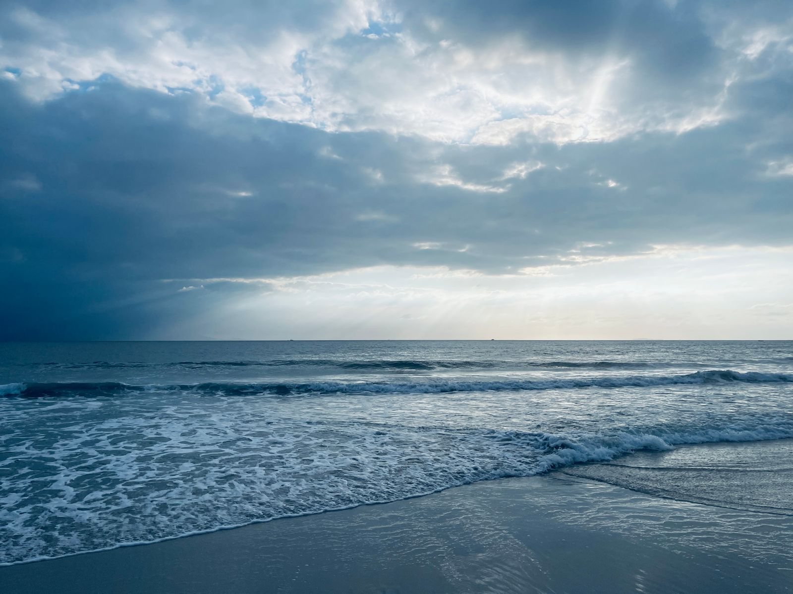 Gentle waves rolling onto a sandy beach under a cloudy sky with sunbeams over the ocean