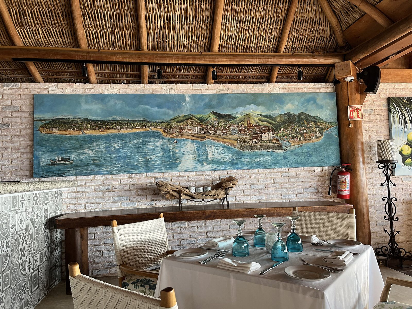 Table with a white tablecloth and blue hand-blown glassware under a palapa roof, with a mural of Puerto Vallarta and the bay on the wall