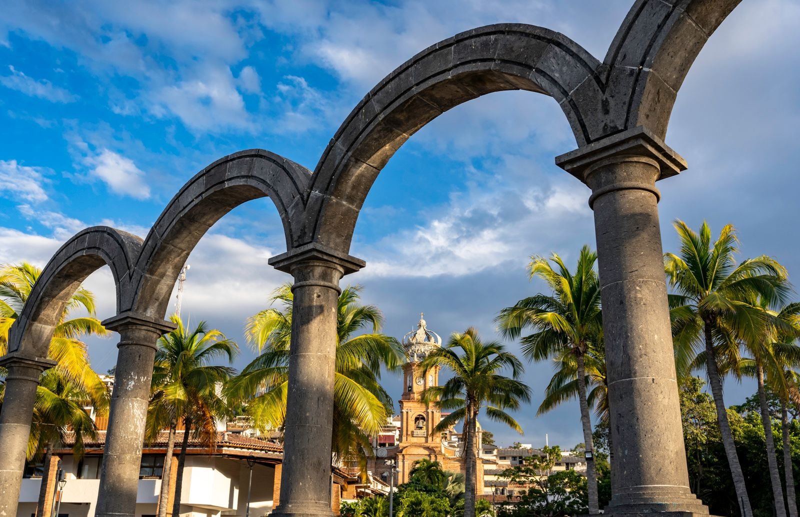 Gray stone arches of the Puerto Vallarta Malecón framing the crowned tower of the Church of Our Lady of Guadalupe among palm trees and tile roofs