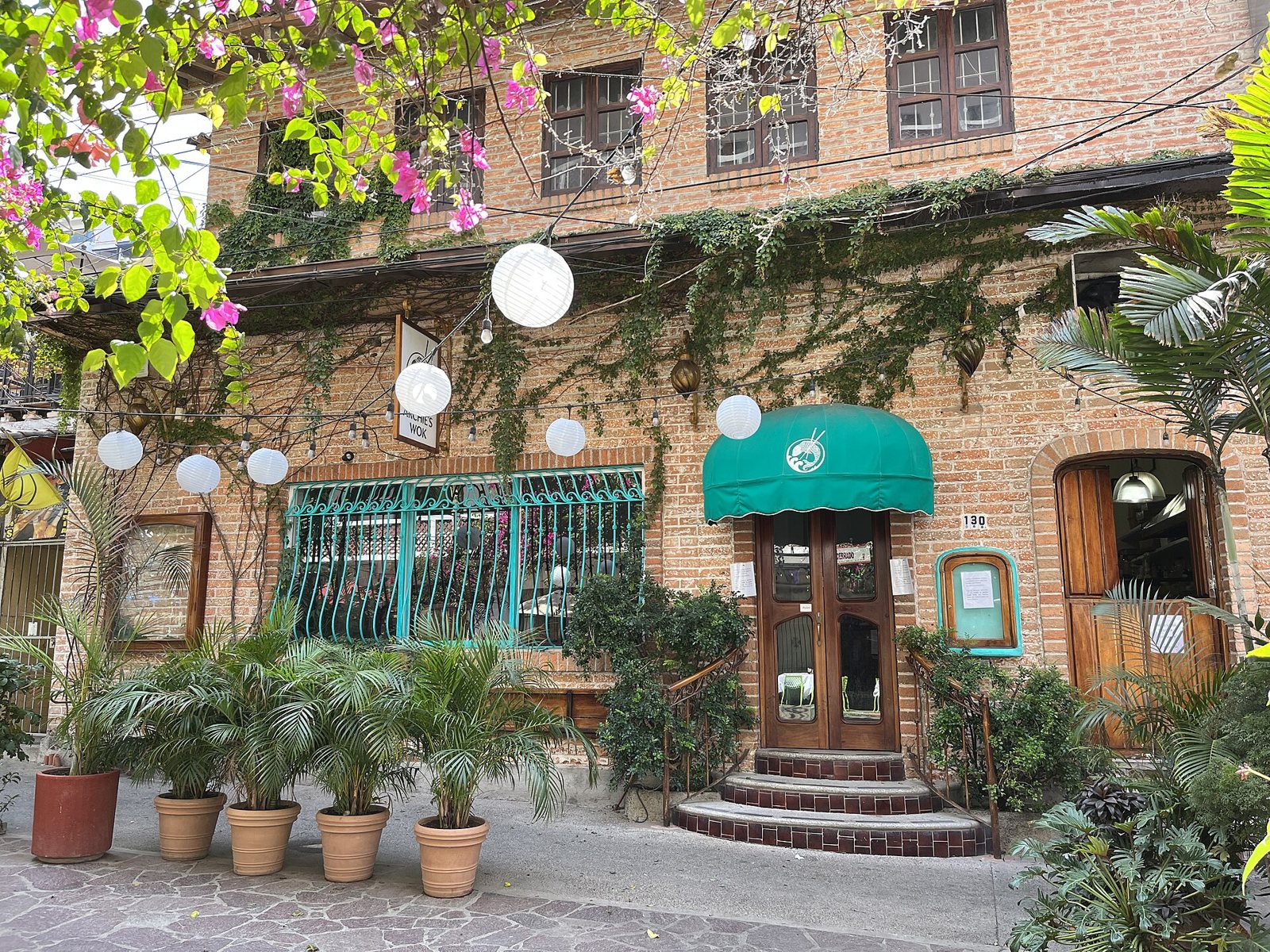 Vine-covered brick facade of Archie's Wok with a green awning, white paper lanterns, bougainvillea and potted palms