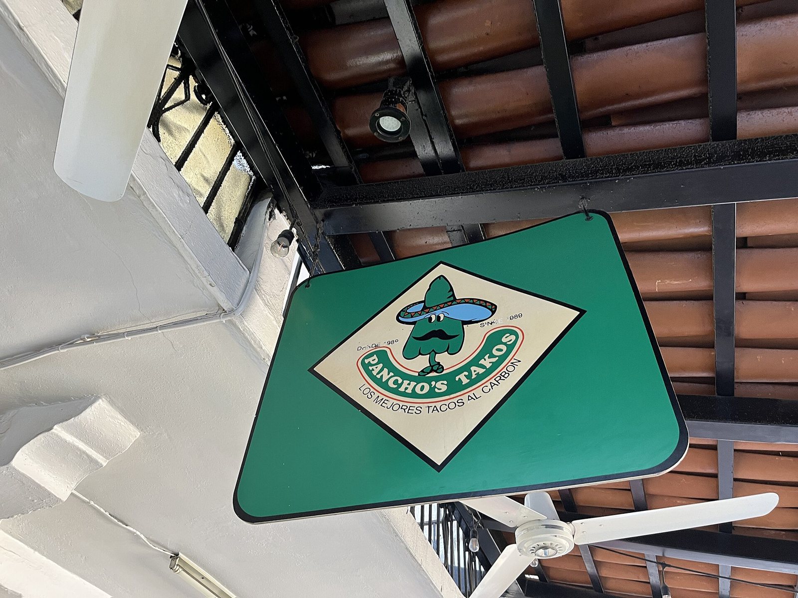 Green hanging sign for Pancho's Takos reading "Los mejores tacos al carbón" under a clay-tile roof