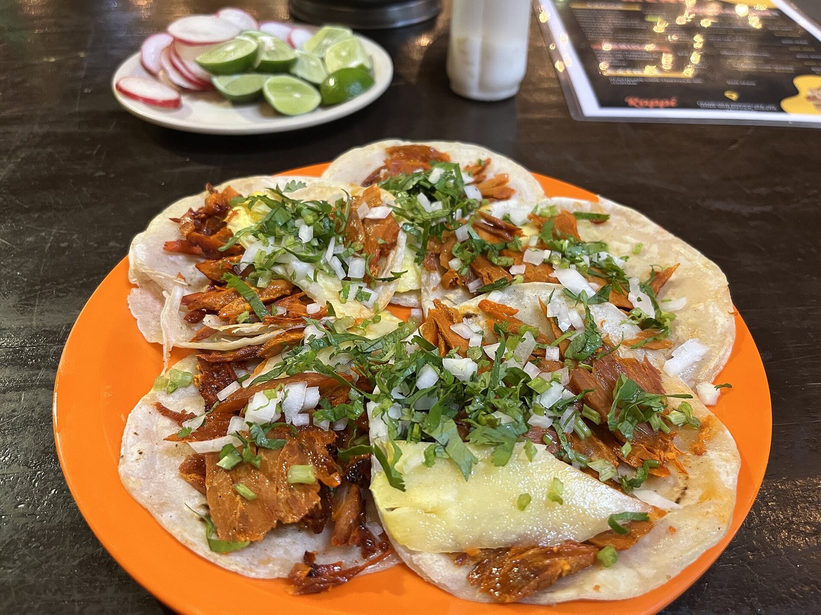 Orange plate with six al pastor tacos topped with pineapple, onion and cilantro, with limes and radishes in the background