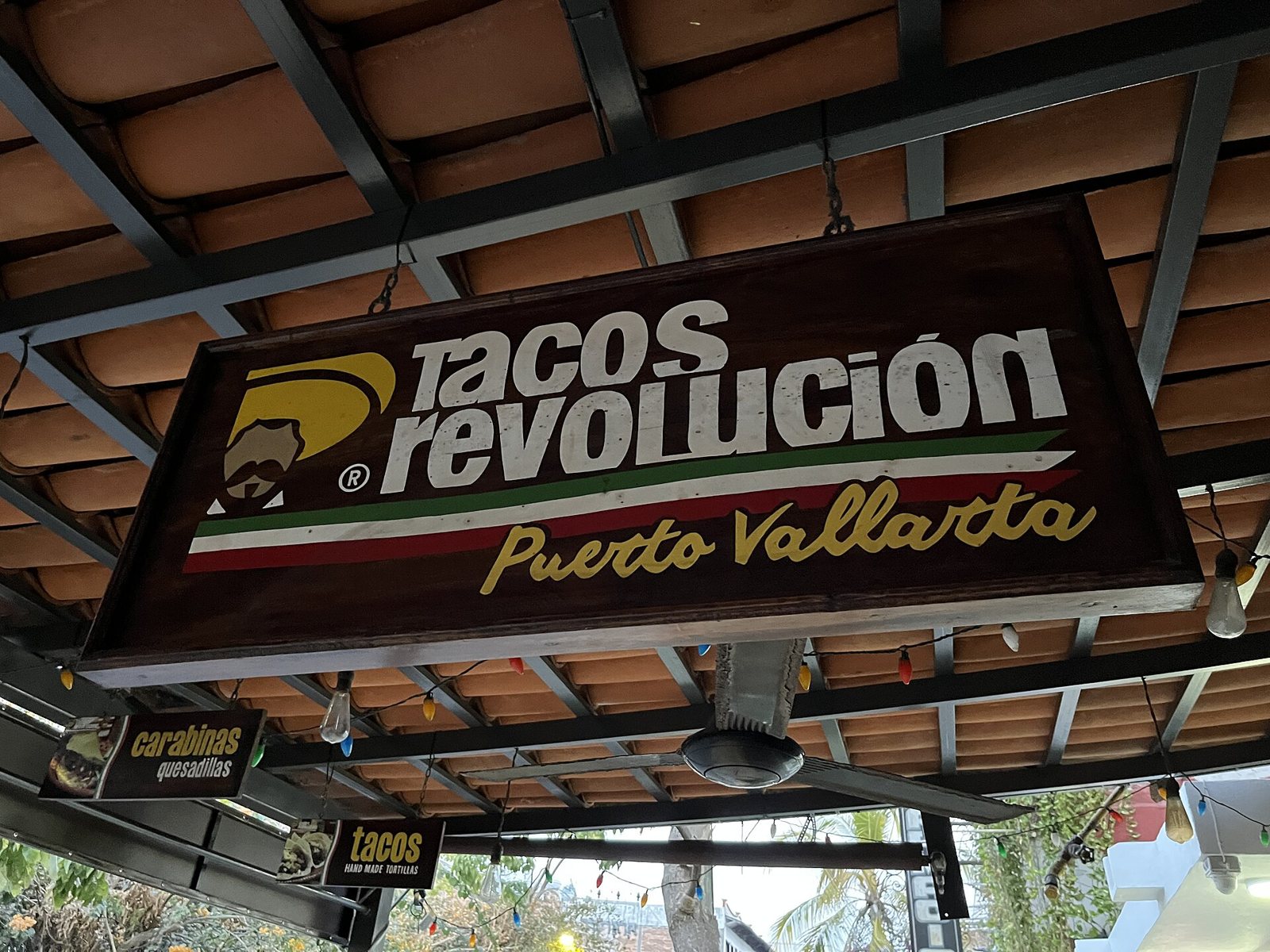 Wooden Tacos Revolución Puerto Vallarta sign with a green, white and red stripe, hanging under a tile roof strung with lights