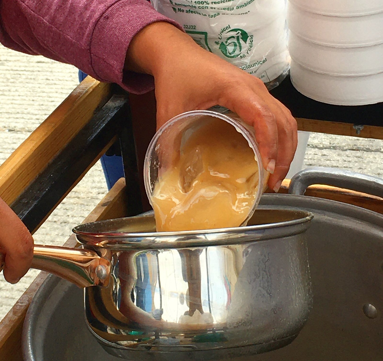 Hands pouring thick tejuino from a cup into a metal pot at a street stand