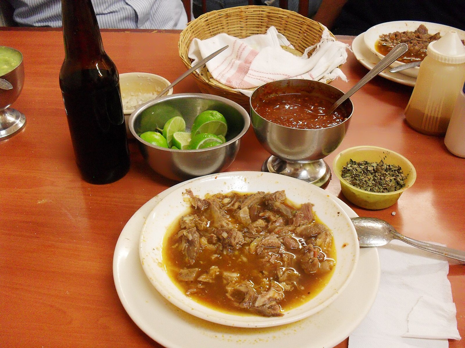 Plate of birria on a table with limes, red salsa, dried oregano, onion and a basket of tortillas