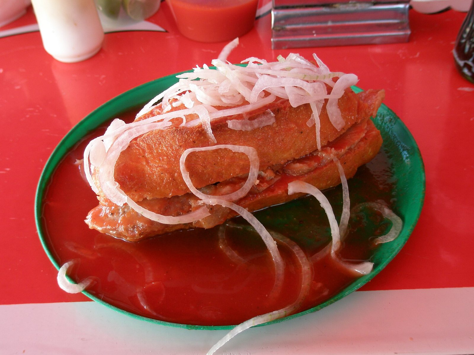 Torta ahogada sandwich drowned in red tomato sauce on a green plate, topped with sliced onion