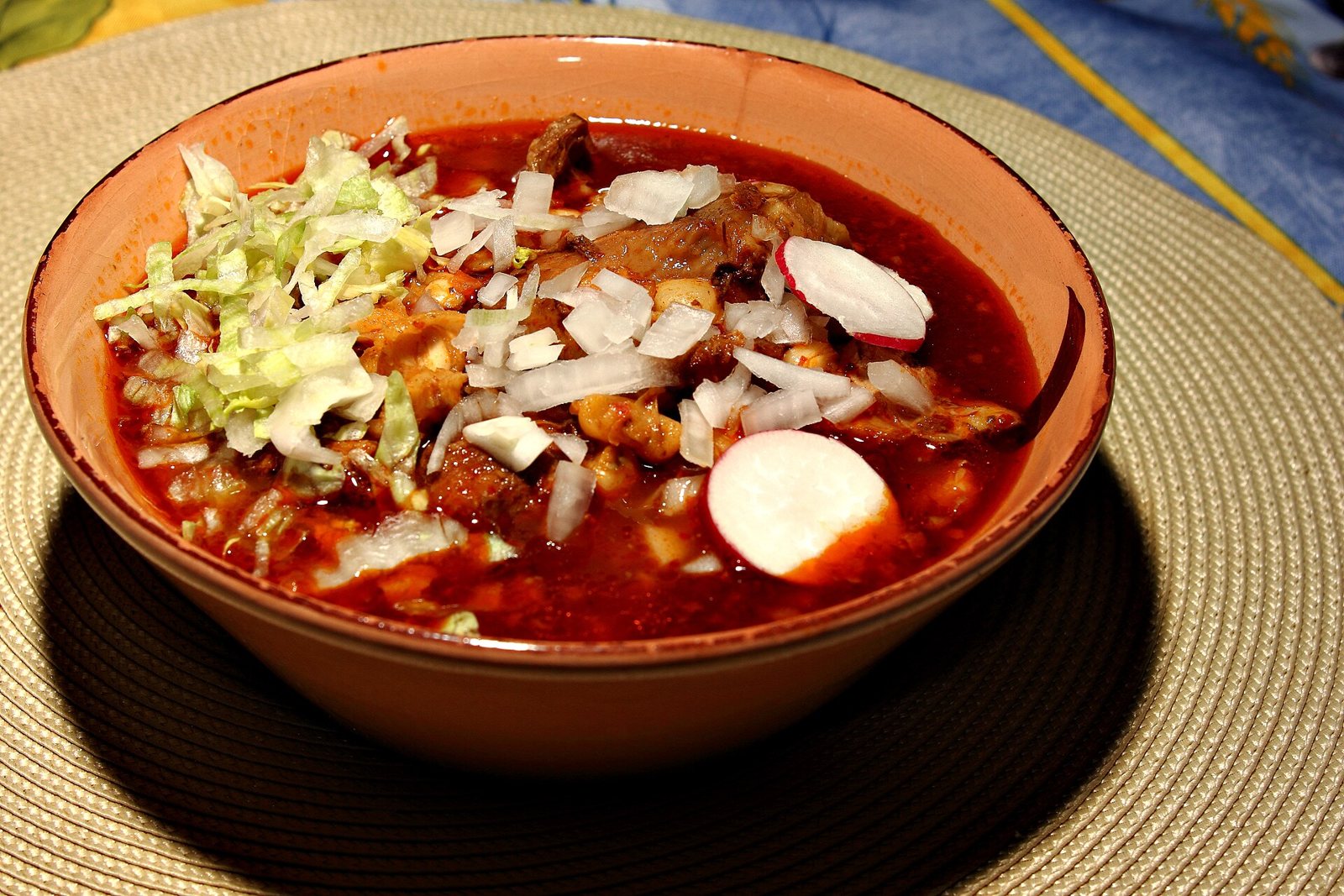 Bowl of red pozole with pork and hominy, topped with shredded lettuce, chopped onion and radish slices
