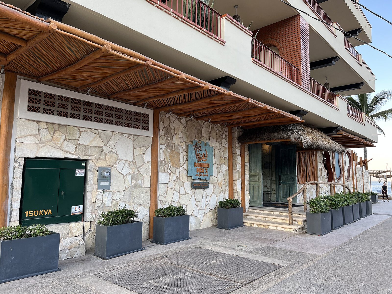 Entrance to Daiquiri Dick's on Olas Altas street, with a stone wall, palm-thatch awning, planters and the ocean in the background