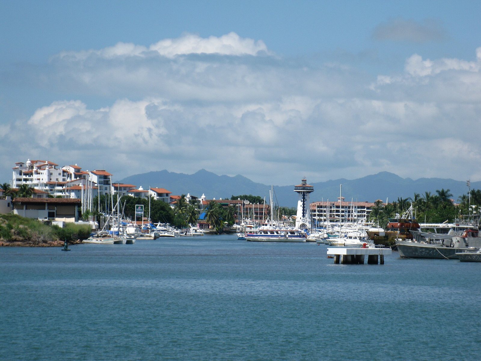 Marina Vallarta seen from the water: sailboats and yachts, white buildings, the lighthouse tower in the middle and the Sierra Madre behind