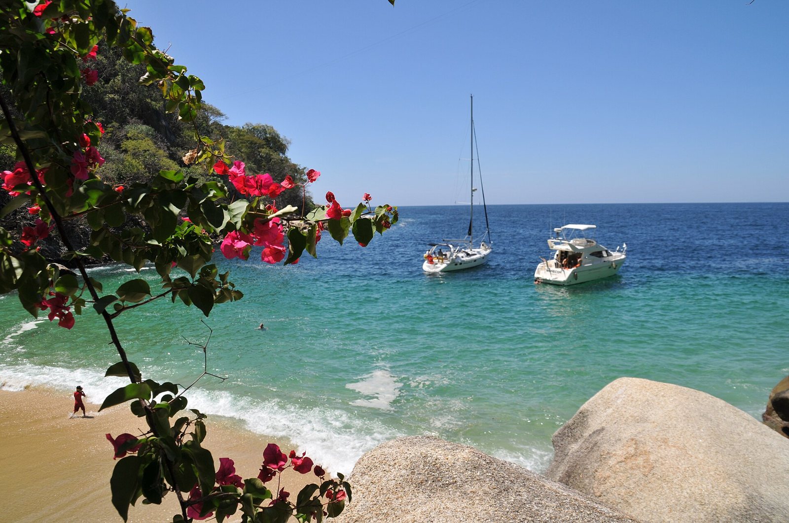 Golden-sand cove with turquoise water and two anchored boats, framed by bougainvillea flowers and granite boulders